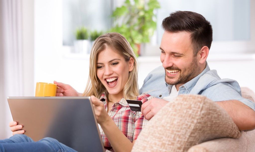 A couple sits on the sofa, happily shopping on their laptops, holding a hot drink and a credit card in their hands, showcasing a joyful online shopping scene.