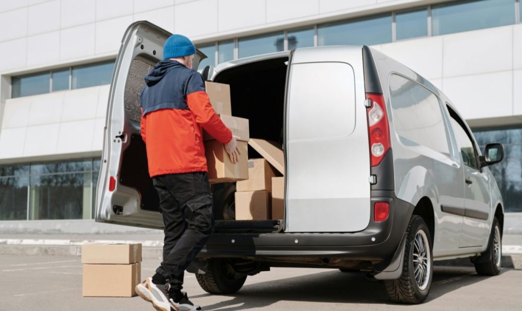 A person wearing a combination of red and black jackets is loading cardboard boxes into the back of a silver truck.