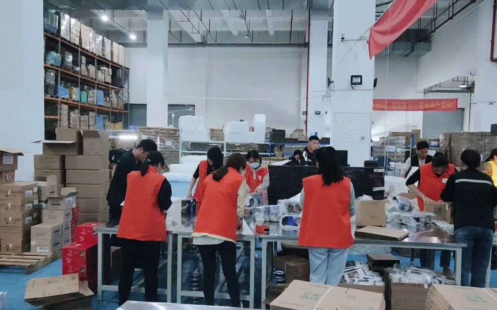 An image of the Runto packing warehouse, showing a dynamic scene where workers are engaged in packing products for shipment. Several individuals in orange vests are collaborating at tables, surrounded by various boxes and products, illustrating a busy and organized packing environment.