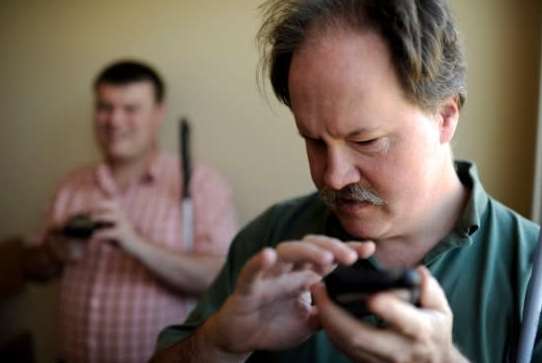 A photograph showing two men, one in the foreground focusing intently on a mobile device in his hands. The man appears to be visually impaired, using tactile feedback to navigate the phone. In the background, another man is also present, holding a mobile device. The setting suggests an environment where assistive technology is utilized to enhance communication and accessibility.