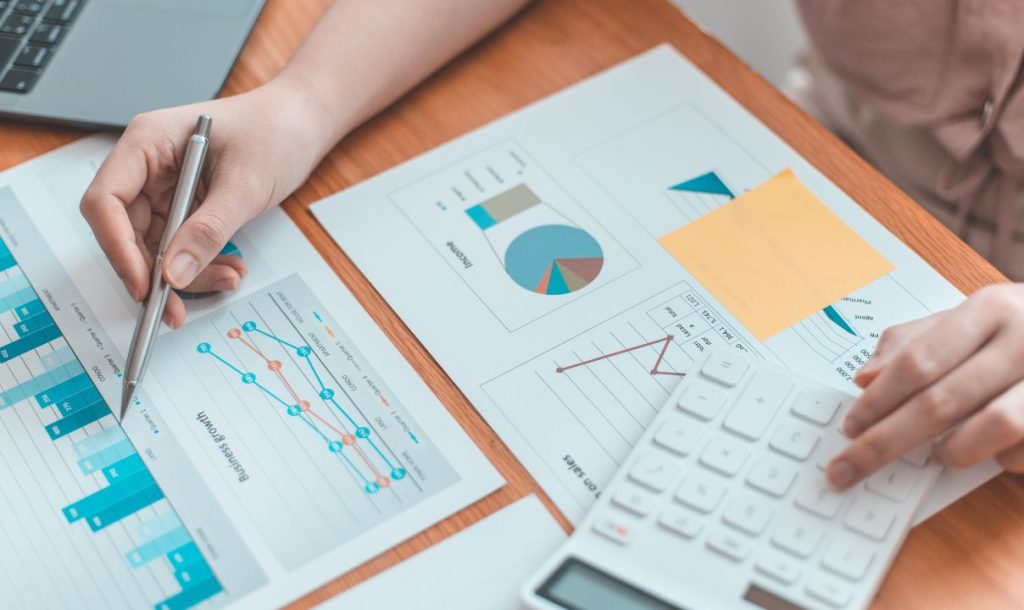 A close-up of a person's hand holding a pen over various financial charts and graphs on paper, with a calculator nearby. The workspace features organized data visualizations, emphasizing a focus on analysis and decision-making in a professional setting.