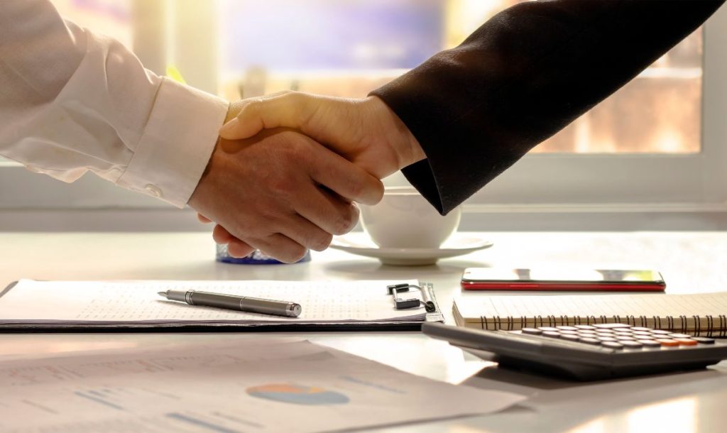 Two individuals are shaking hands in a business setting, symbolizing a successful agreement. A pen, notebook, and calculator are visible on the table, along with documents featuring charts, suggesting a professional discussion or negotiation has taken place.