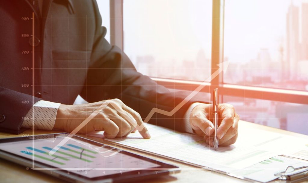 A businessman analyzing financial charts and graphs while writing notes, with a tablet displaying data trends next to him. An upward trend graph is superimposed on the image, highlighting growth.