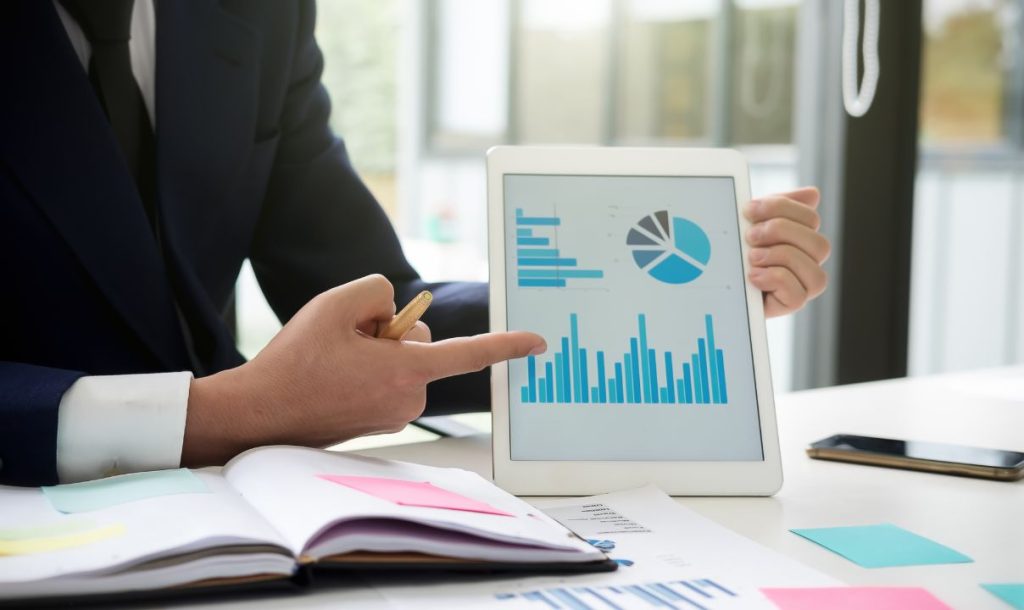  A businessman in a suit points at a tablet displaying colorful graphs and charts, illustrating data analysis. A notebook with sticky notes and documents is placed on the table, indicating a collaborative or presentation setting. The background shows a bright