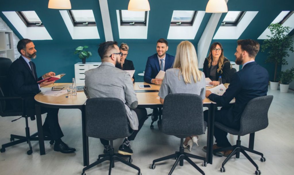 A group of professionals sitting around a conference table in a modern office, engaged in a business discussion. The team consists of men and women dressed in formal business attire, with documents and tablets in front of them. The room features natural light coming through skylights and plants for a fresh atmosphere.