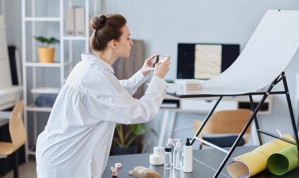 A woman in a white shirt focuses intently while photographing beauty products arranged on a tabletop. She is positioned in a well-lit studio with shelves in the background and various items, including containers and colored paper rolls, indicating a creative workspace for product styling and photography.