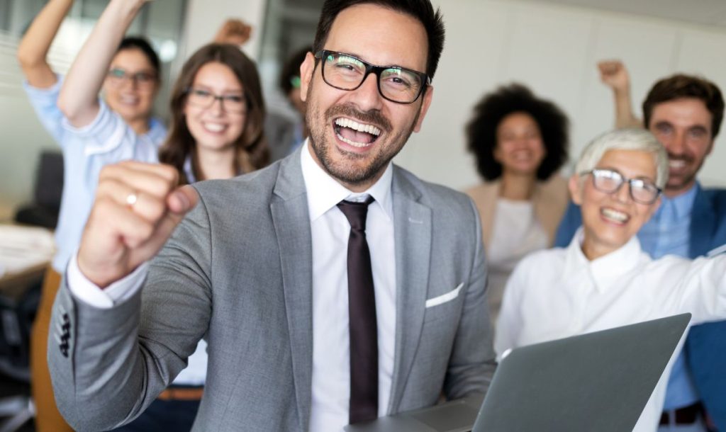A group of happy colleagues celebrates success in an office setting. The man in a gray suit in the foreground is smiling widely, raising his fist in triumph while holding a laptop. Behind him, a diverse group of teammates, all smiling and cheering, express joy and excitement, capturing a moment of teamwork and achievement.