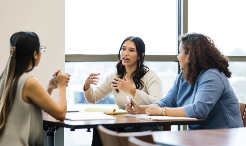 Three women engaged in a discussion during a business meeting, with one actively sharing ideas while the others listen attentively. They are seated around a table with notepads and a large window in the background.
