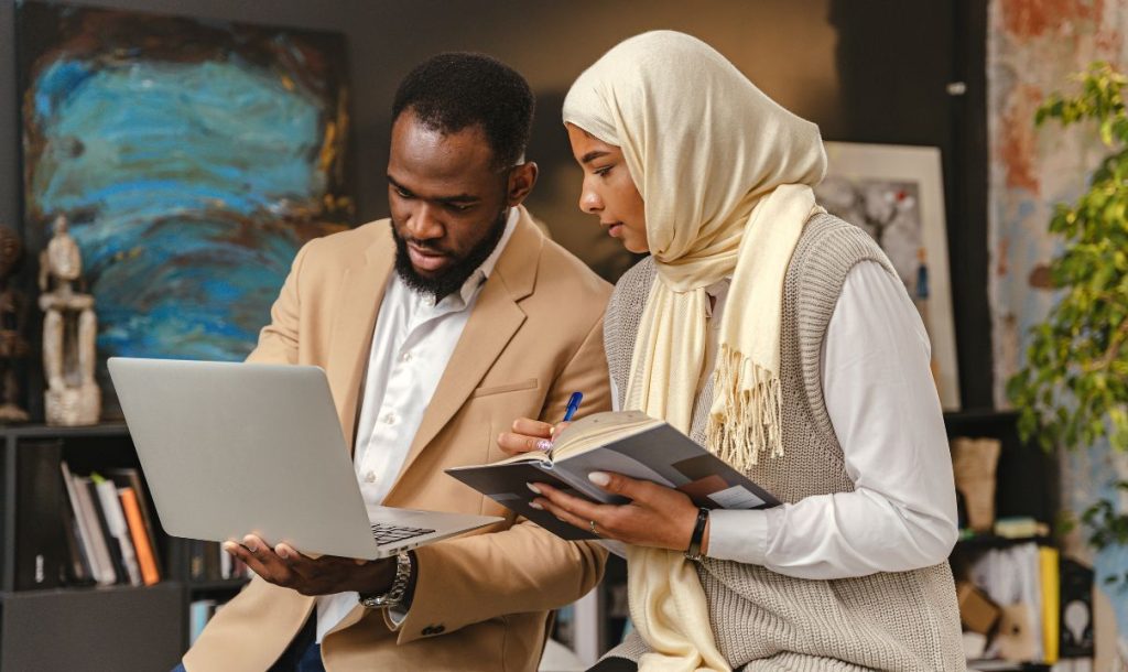 A man and a woman are engaged in a discussion during a study session, with the man holding a laptop and the woman taking notes in a notebook. The woman is wearing a hijab and a cozy sweater, as they both focus on the task at hand in a modern workspace.