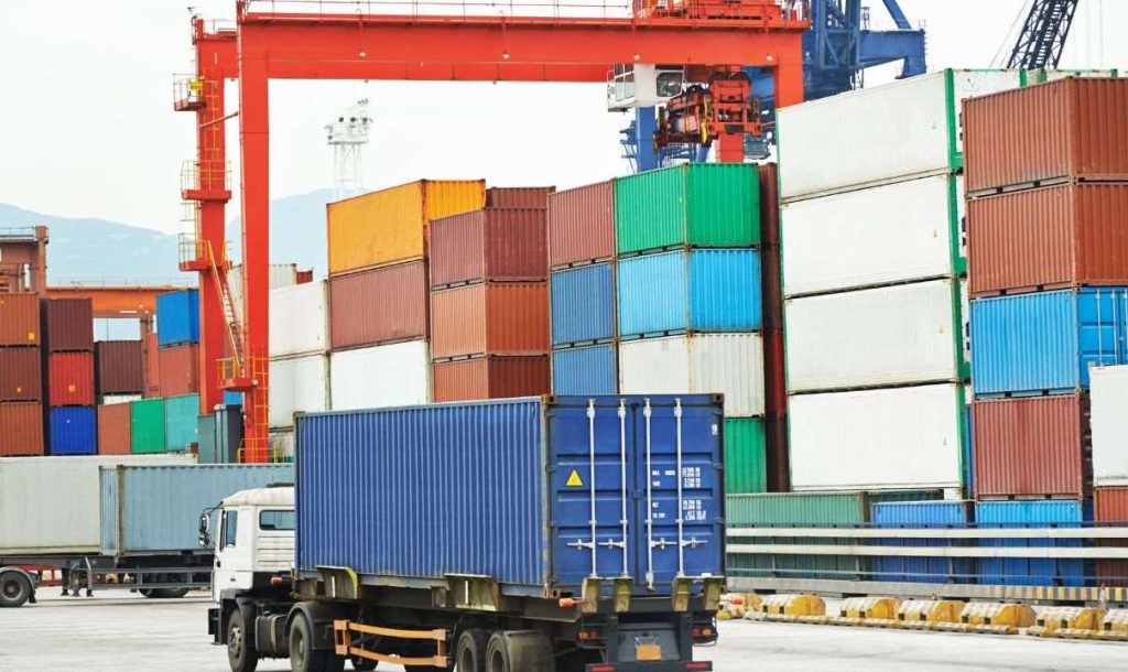 A busy shipping container yard featuring stacked cargo containers of various colors. A truck is positioned for loading or unloading beside the containers. A crane is visible in the background, indicating ongoing port operations.