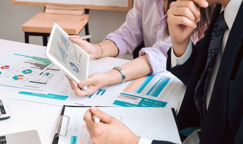 Two professionals are engaged in a discussion over charts and financial reports on a table. One person is holding a tablet displaying graphs, while the other appears to be thoughtfully analyzing the information, indicating a collaborative business planning session.