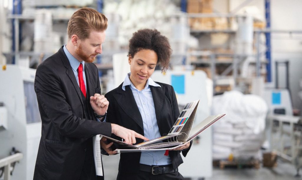 Two professionals, a man and a woman, are reviewing a product design book in a manufacturing facility. The environment features machinery in the background, indicating a business setting focused on production and design collaboration.