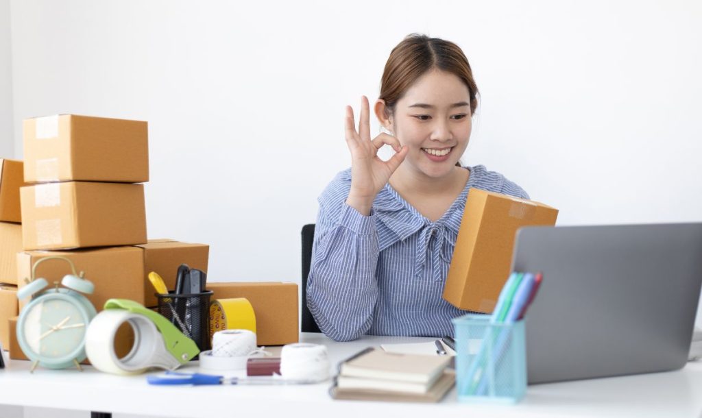 A young woman smiling and showing an "okay" hand gesture while holding a cardboard box in front of her laptop. She is surrounded by several stacked boxes and various office supplies, including tape and pens, on a desk. The setting suggests an e-commerce or dropshipping business environment.