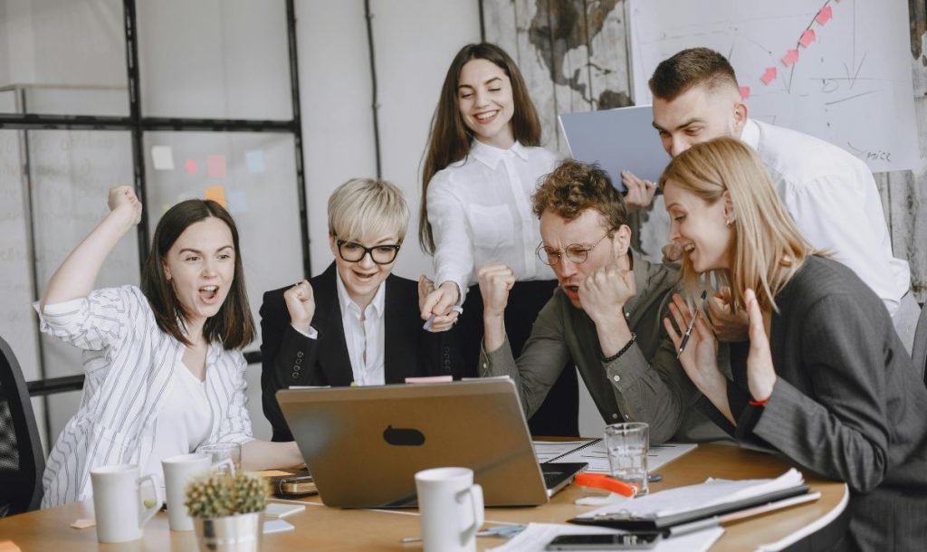 A group of six colleagues celebrating in an office, showing excitement and cheering in front of a laptop. The team is expressing joy and enthusiasm, with colorful sticky notes and a whiteboard in the background.