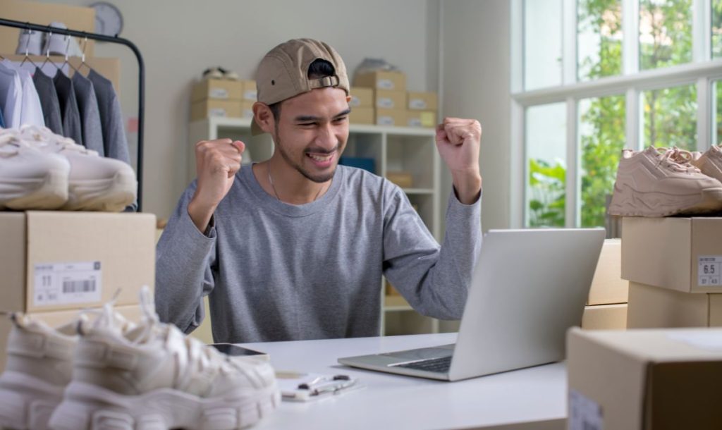 A young man is celebrating triumphantly while sitting at a desk with a laptop, surrounded by boxes of shoes. His enthusiastic expression suggests a successful purchase or achievement related to online shopping.