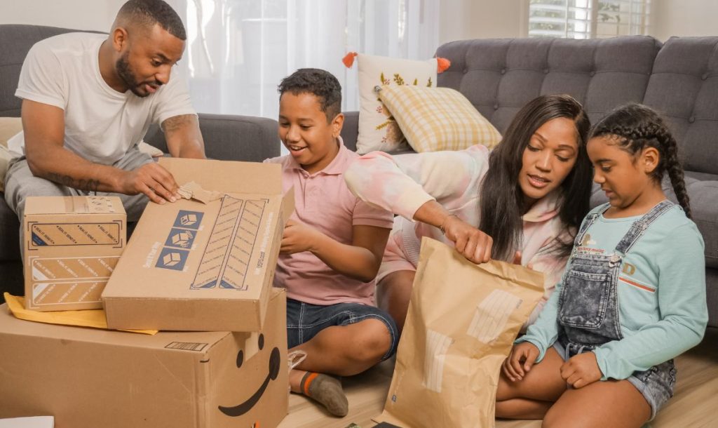 A family is sitting on the floor in a living room, joyfully unboxing packages together. The parents and two children are engaged, with various cardboard boxes and envelopes scattered around them.