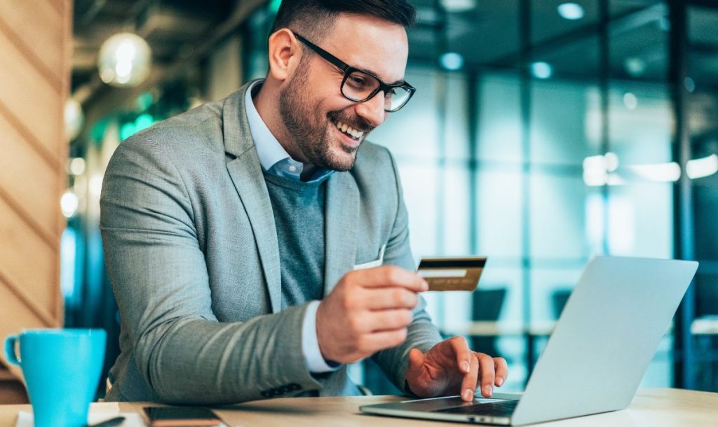  A man in a gray suit and glasses is smiling while holding a credit card and using a laptop. He is sitting at a table in a modern office environment, with a blue coffee cup and a smartphone visible in the foreground, capturing a moment of joy in online shopping.