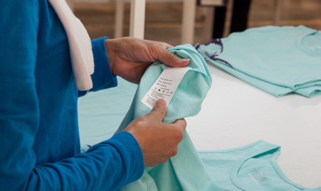 A close-up of hands holding a folded light blue t-shirt while examining the care label. In the background, additional shirts are visible, indicating a setting focused on clothing handling and quality control.