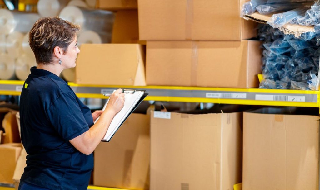 A woman in a dark polo shirt is checking inventory in a warehouse, using a clipboard and pen while standing in front of shelves filled with cardboard boxes and packaging materials.