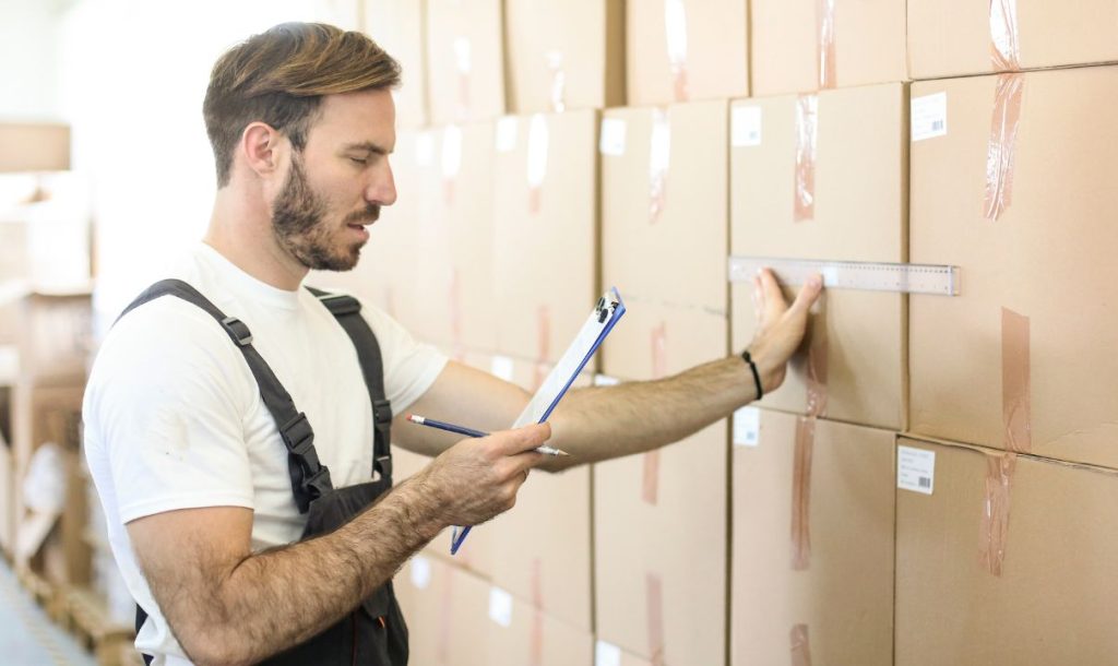 A man is measuring the height of stacked boxes in a warehouse while holding a clipboard and pencil. The image illustrates the process of inventory checking and organization in a storage environment.