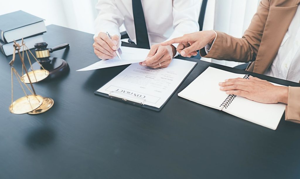 A close-up view of a legal consultation where one individual is reviewing a contract while the other points out key details. On the table, there are legal books, a gavel, scales of justice, and a notebook, indicating a professional setting focused on legal matters and agreements.