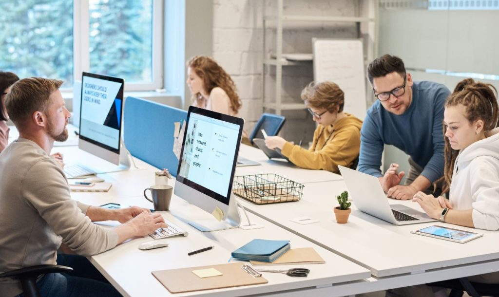 A group of people collaborate in a modern office, sitting at a desk to work, using computers and laptops, creating an atmosphere of focus and discussion.