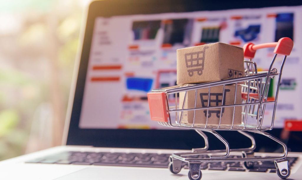A miniature shopping cart loaded with small cardboard boxes sits on a laptop keyboard, while an online store interface is visible in the background, showcasing various products.