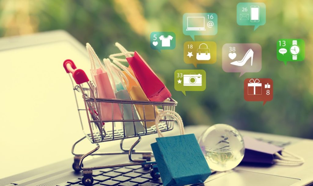 A shopping cart filled with colorful bags placed on a laptop keyboard, surrounded by various digital icons representing online shopping categories and notifications. The background features soft greenery, suggesting a fresh shopping experience.
