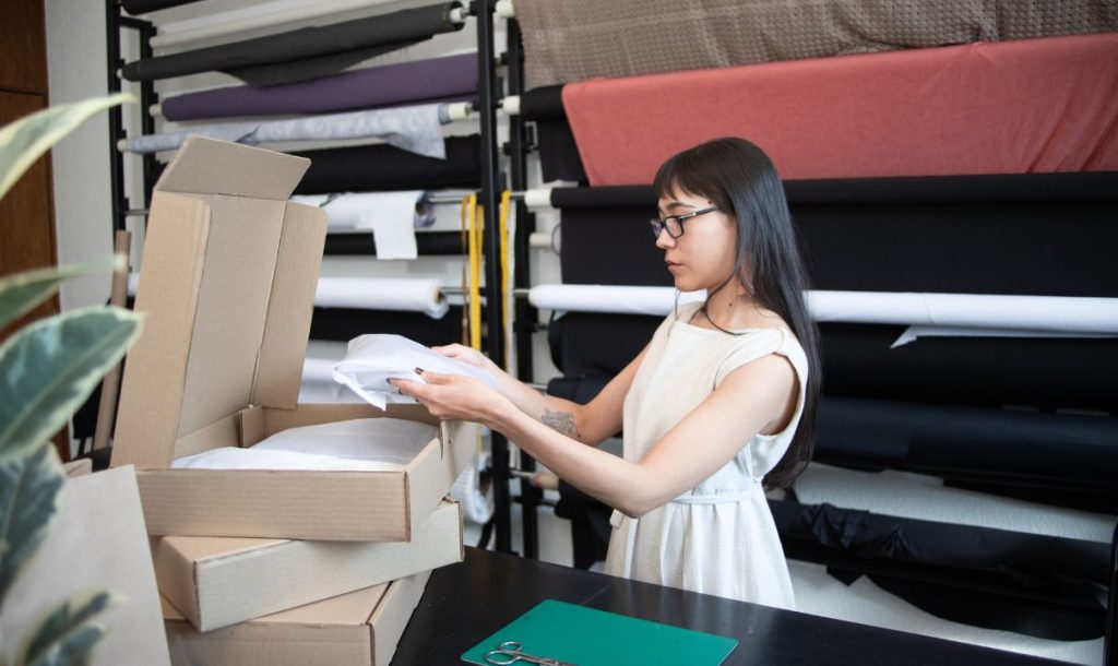 A woman with glasses is examining folded fabrics while surrounded by open boxes in a workspace filled with rolls of material. The setting reflects a creative environment, emphasizing the process of organizing and preparing items for sale or shipment.
