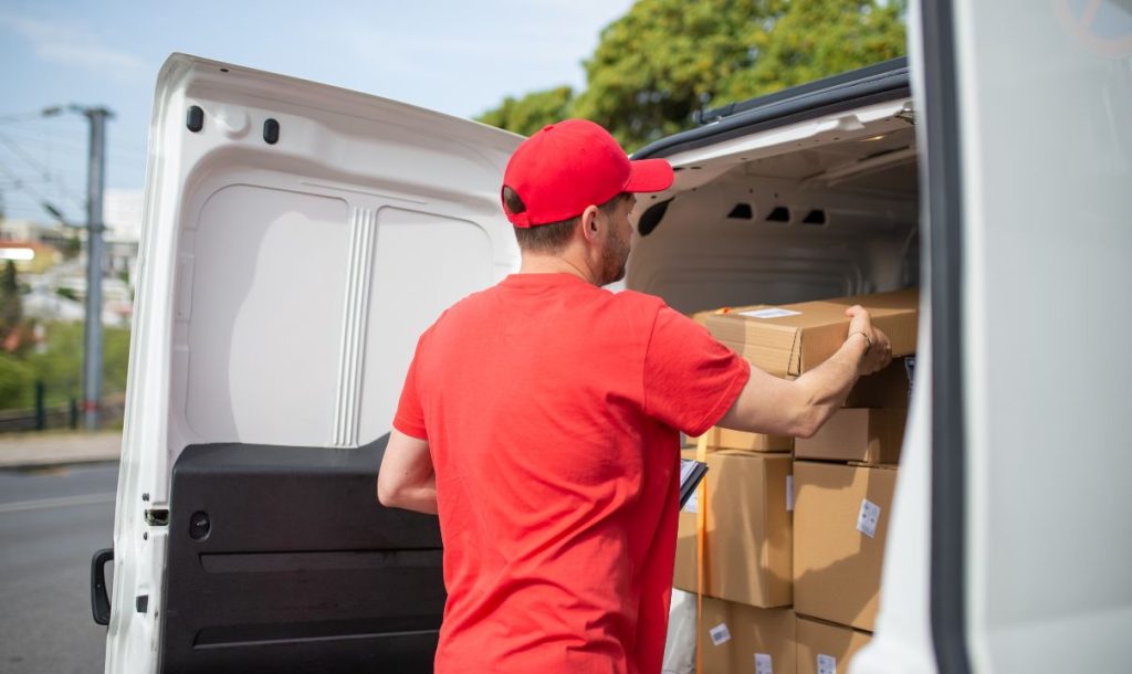A delivery person in a red shirt and cap is loading boxes into a white van, with packages stacked inside the vehicle, set against a backdrop of trees and a street.