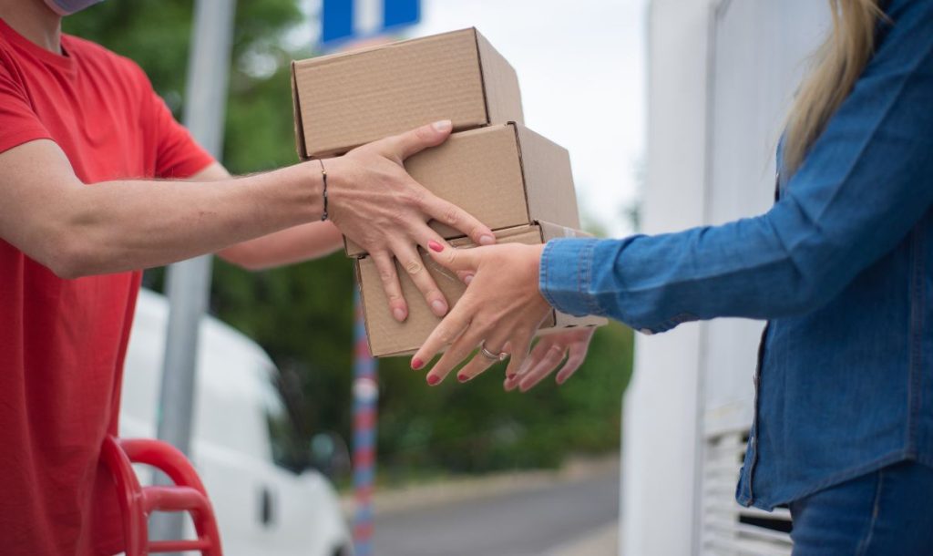 A delivery person wearing a mask is handing over three cardboard boxes to a female recipient, with both hands reaching out to take or receive the packages. A blurred background features a street scene.