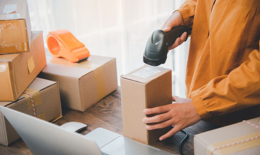 A person is scanning a shipping label on a cardboard box using a handheld scanner. The workspace is cluttered with packed boxes and packing tape, with a laptop visible in the background, indicating a busy shipping or e-commerce environment.