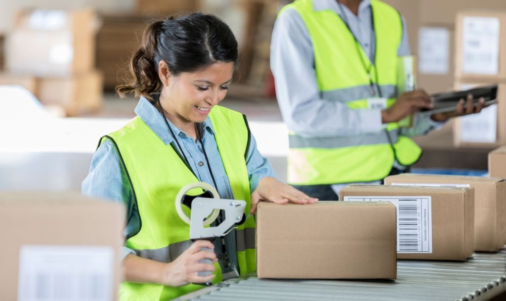A woman in a neon safety vest is working at a packing station, smiling as she applies a label to a cardboard box. In the background, a colleague is using a tablet, contributing to the efficient workflow in a busy warehouse environment.