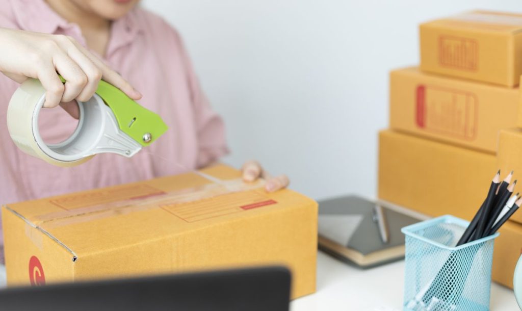 A close-up image of a person using a tape dispenser to seal a cardboard box. The background includes other stacked boxes and office supplies, creating a workspace atmosphere focused on packaging and shipping.