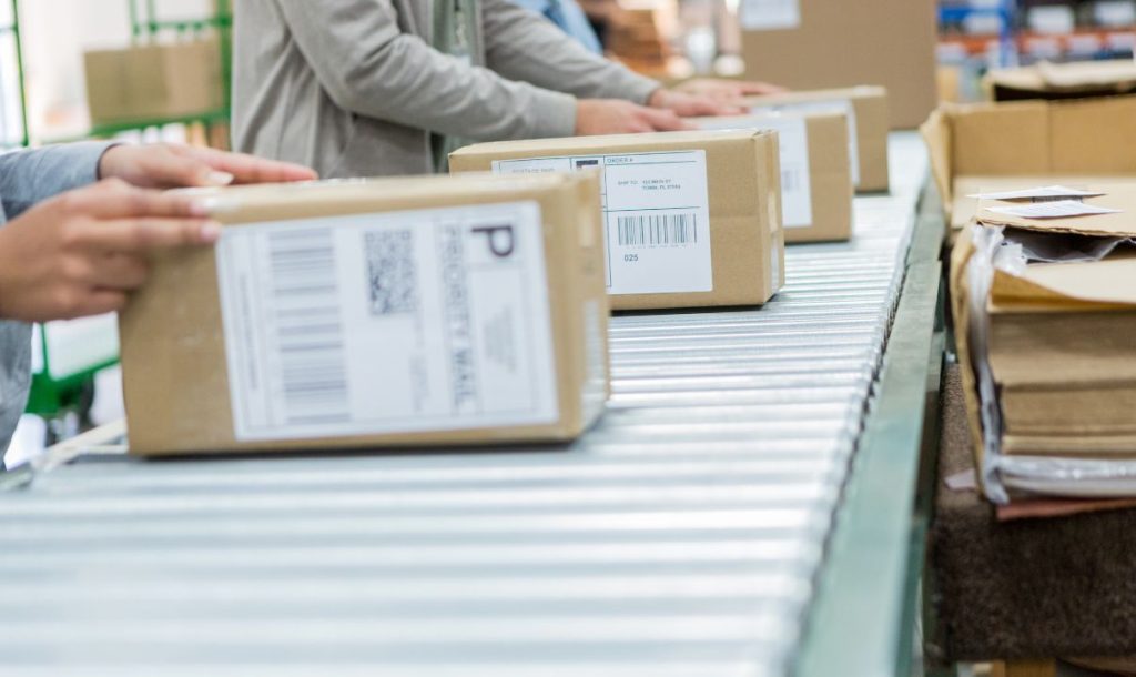 A close-up view of a packing line in a warehouse where multiple hands are seen handling packages with shipping labels. The image depicts the operational workflow involved in preparing orders for shipment.