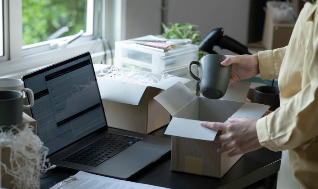  A person is packing mugs into a cardboard box at a desk with a laptop open, surrounded by packing materials. The scene shows a workspace filled with boxes and items, indicating an order fulfillment process.