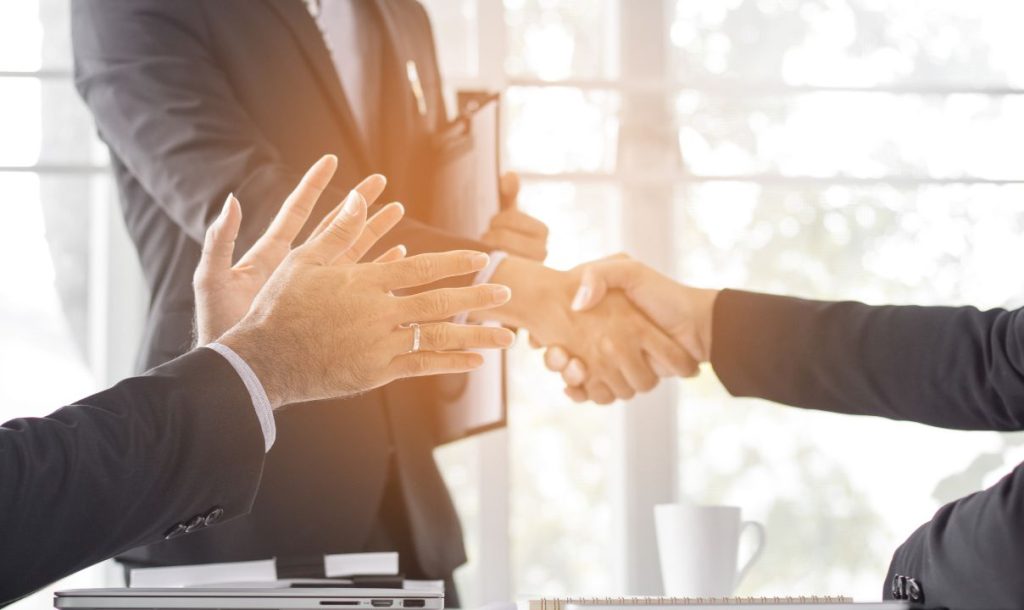 Two business people shook hands at the conference table, one holding a folder and the other with both hands forward. There is a laptop and a cup of coffee on the table.