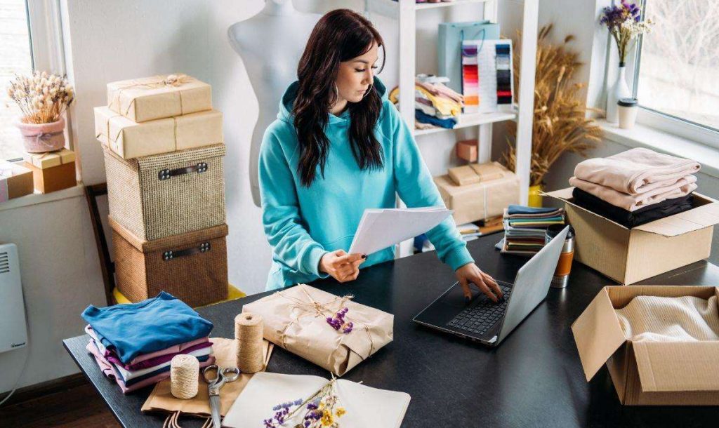 A woman in a blue hoodie examines paperwork while working on a laptop in a creative workspace filled with fabrics, gift boxes, and craft supplies.