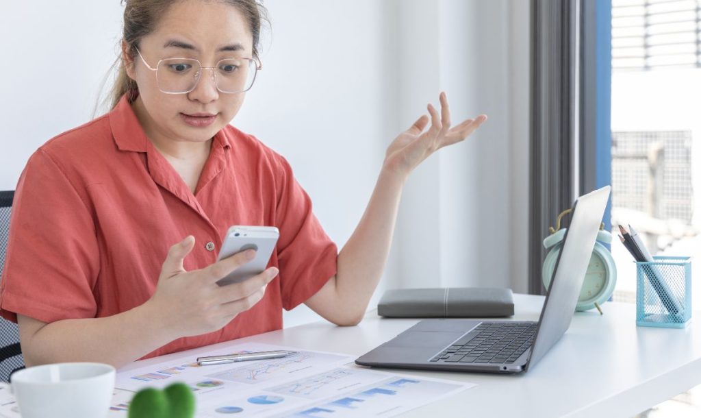 A woman with glasses looks astonished while holding a smartphone in one hand and gesturing with the other in a bright office environment. A laptop, documents with charts, and office supplies are visible on the desk, illustrating a moment of surprise or confusion during her work.