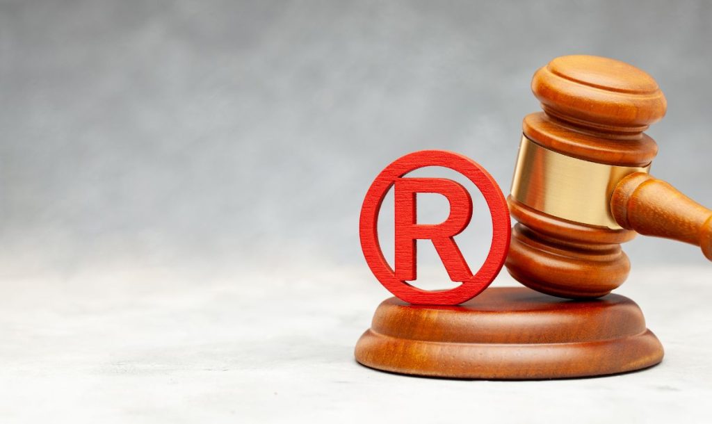 A wooden gavel placed beside a red registered trademark symbol on a textured background. The image symbolizes legal authority and the importance of trademark protection in business and intellectual property rights.