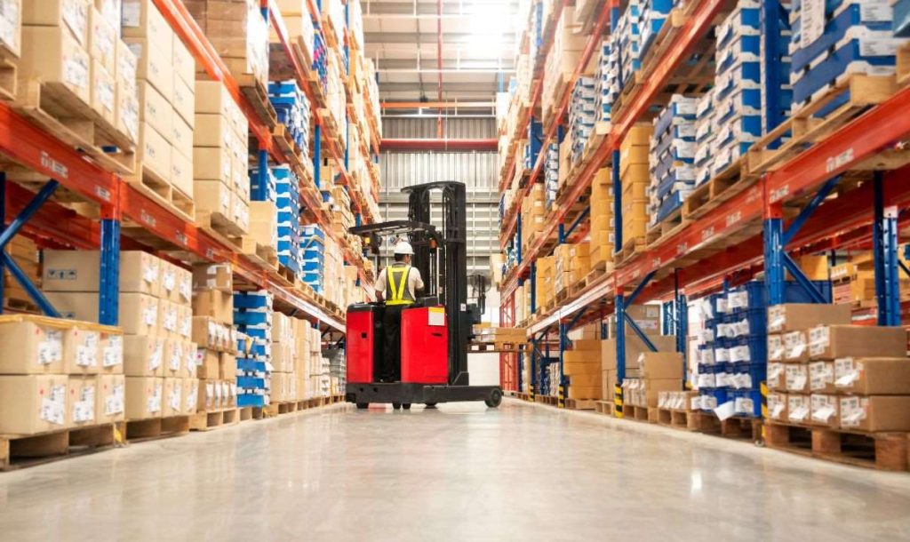 A forklift operator is maneuvering through a warehouse filled with tall shelves stacked with cardboard boxes. The image captures the organized layout of the storage facility, showcasing an efficient logistics environment.