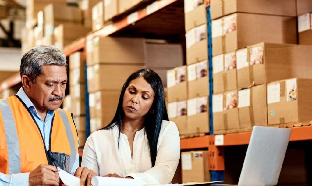 Two warehouse workers, a man in a safety vest and a woman in a white shirt, reviewing documents together in a storage area filled with stacked boxes. A laptop is visible on the table, indicating a focus on logistics and inventory management.