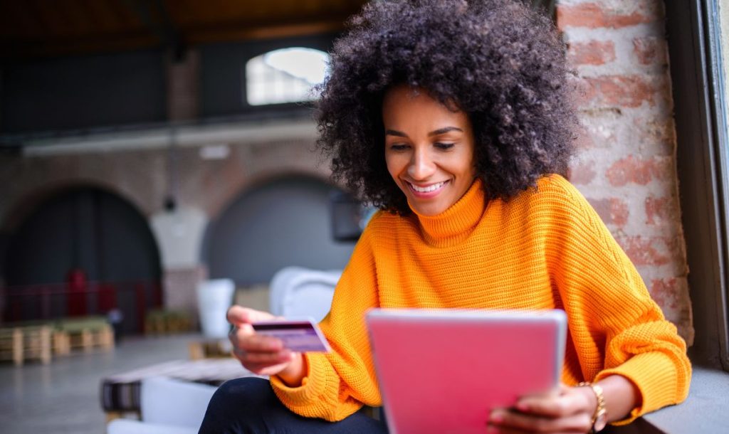 A woman wearing an orange sweater smiled, holding a credit card in her left hand and using a tablet in her right hand, in a comfortable indoor environment.