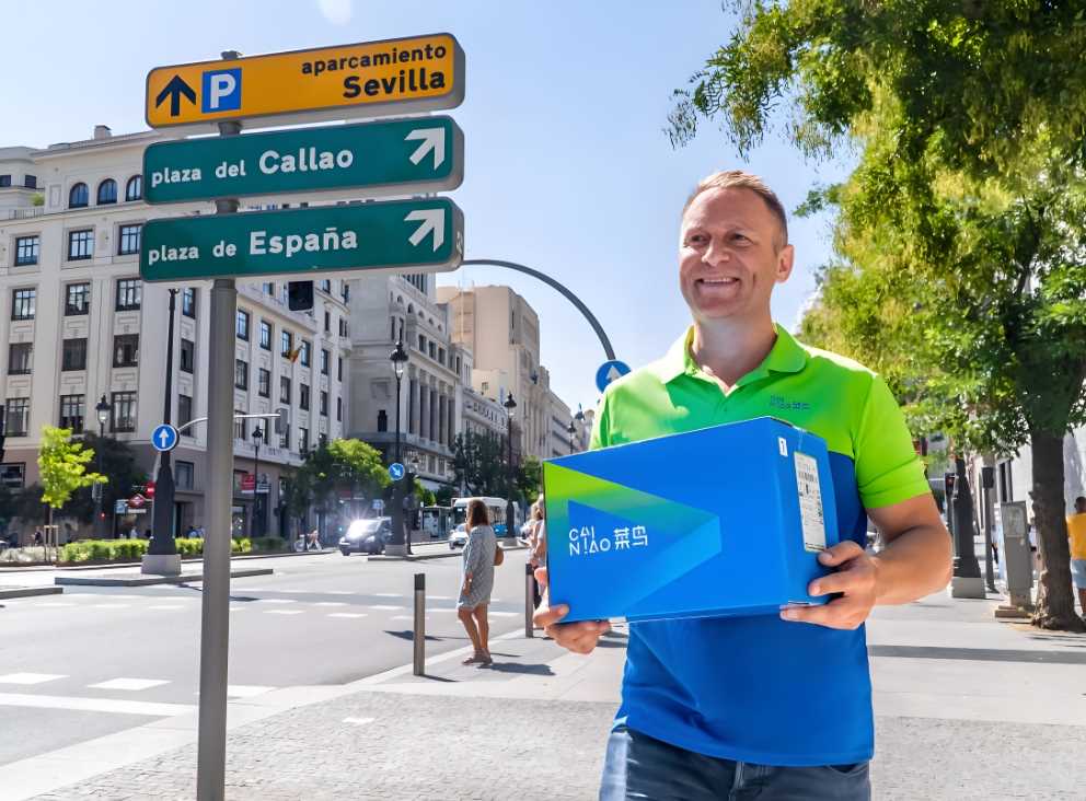 A delivery person wearing a bright green and blue shirt is smiling while holding a Cainiao-branded package. In the background, there are street signs pointing to various locations in Sevilla, including "plaza del Callao" and "plaza de España." The setting is a sunny urban street with pedestrians and trees.
