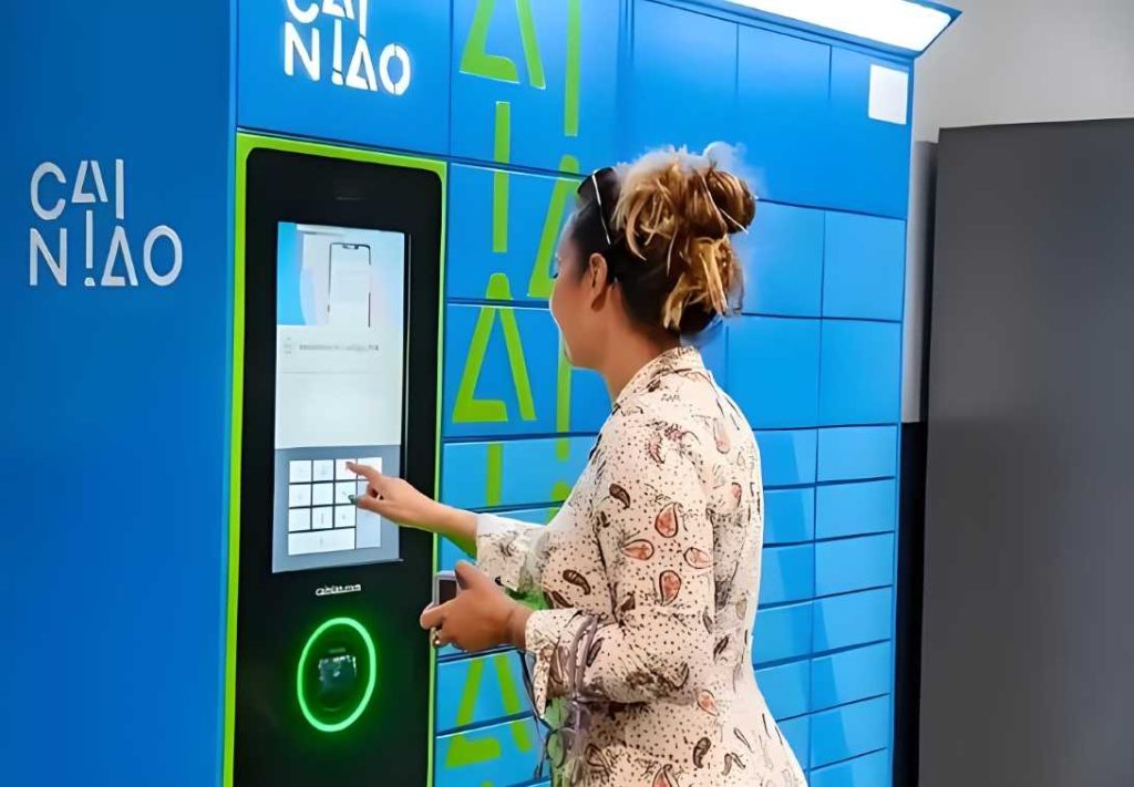 A woman interacts with a digital touchscreen on a Cainiao package locker, entering information to retrieve a package. The locker is bright blue with the Cainiao logo prominently displayed. The setting suggests a modern delivery or collection point.