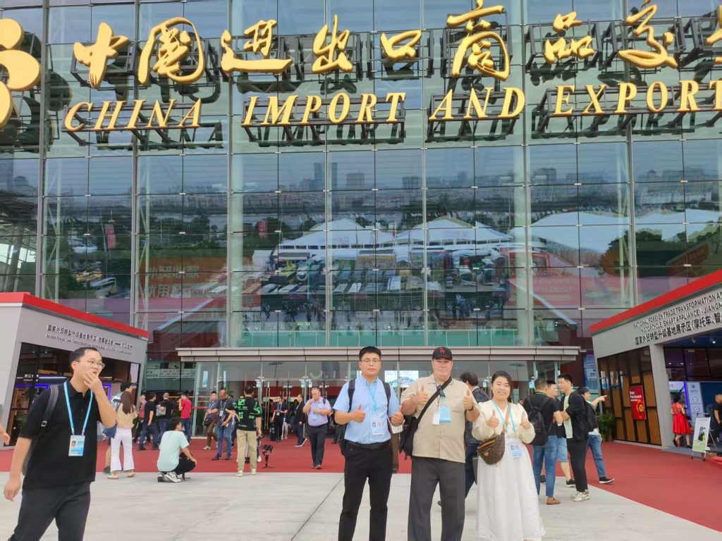 Photo of RuntoAgent representatives and clients standing in front of the China Import and Export Fair building, smiling and giving thumbs up, with the exhibition hall and crowd visible in the background.