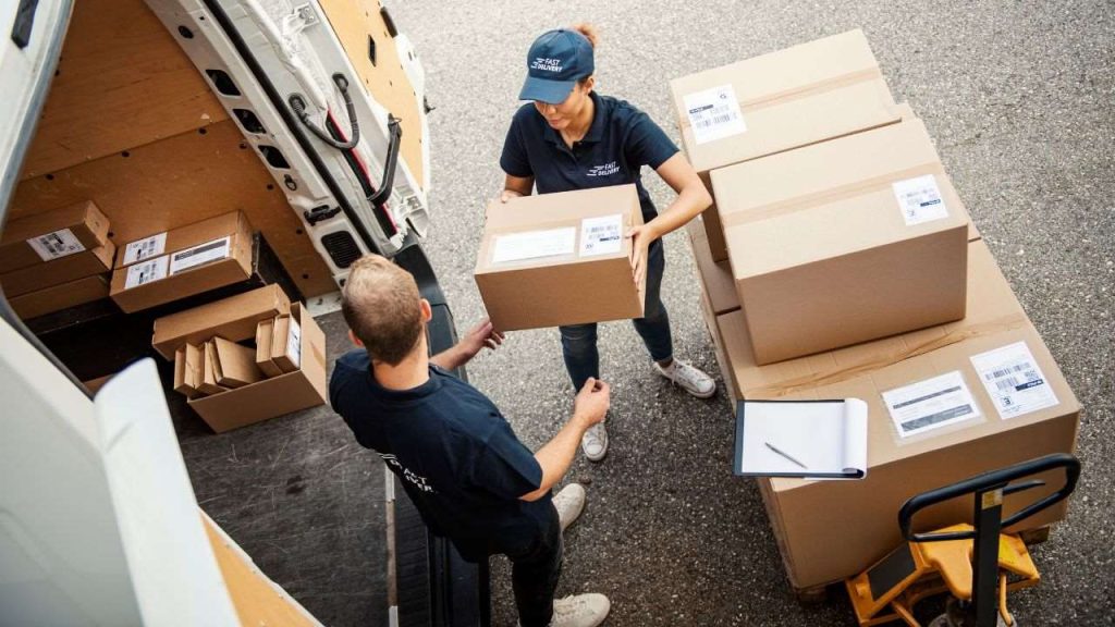 An image depicting a delivery worker passing a parcel to a colleague at the back of a delivery van. Several boxes are organized inside the van, highlighting an efficient loading and unloading process in the logistics operation. The workers are in matching uniforms, emphasizing teamwork and organization.