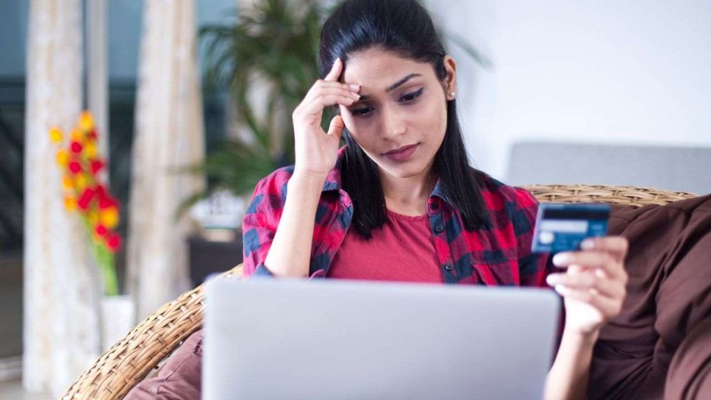 An image of a woman looking concerned as she sits on a couch, holding a credit card in one hand while using a laptop. Her facial expression reflects frustration, likely due to not being able to find information about her online order. The background features plants and a cozy interior setting, emphasizing a typical home environment.