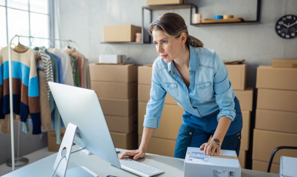 A woman focused on her work at a desk, using a computer while holding a package. She is in an organized workspace filled with clothing displays and cardboard boxes, indicating an e-commerce or retail environment. Her expression shows concentration as she manages inventory or shipping tasks.