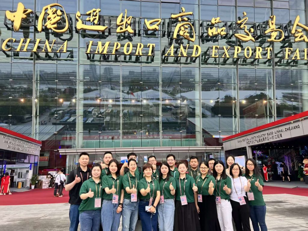 A large group photo of the Runtodropshipping team in front of the China Import and Export Fair building. The team members are wearing matching green shirts and are all smiling while giving thumbs-up gestures. The backdrop features the iconic entrance of the fair, showcasing large signage in both Chinese and English. The vibrant atmosphere indicates a bustling trade event.